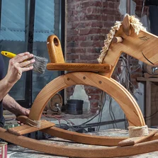 Einfache Verarbeitung - Ein Holzschaukelpferd wird mit einem Pinsel mit Holzlasur Holzschutz transparent gestrichen.